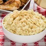 Creamy potato salad in a white bowl with BBQ and pickles on a checkered tablecloth.