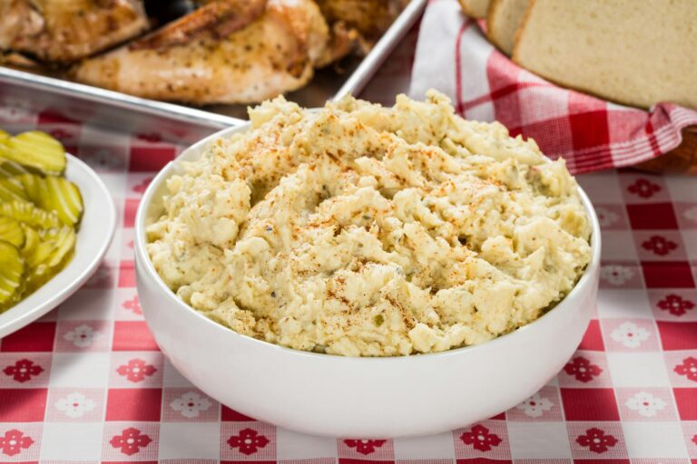 Creamy potato salad in a white bowl with BBQ and pickles on a checkered tablecloth.