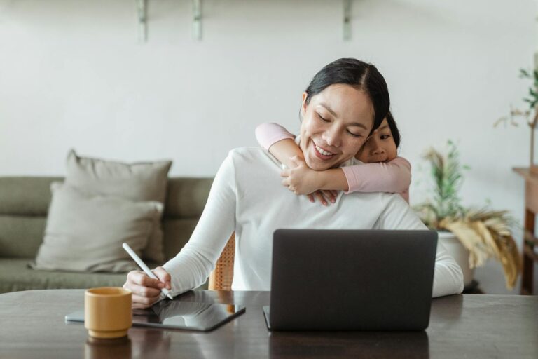 A mother works from home with her daughter embracing her, showing love and family connection.